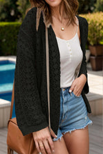 Woman wearing a black openwork open front dropped shoulder cardigan, white tank top, denim shorts, and a brown handbag by the pool.