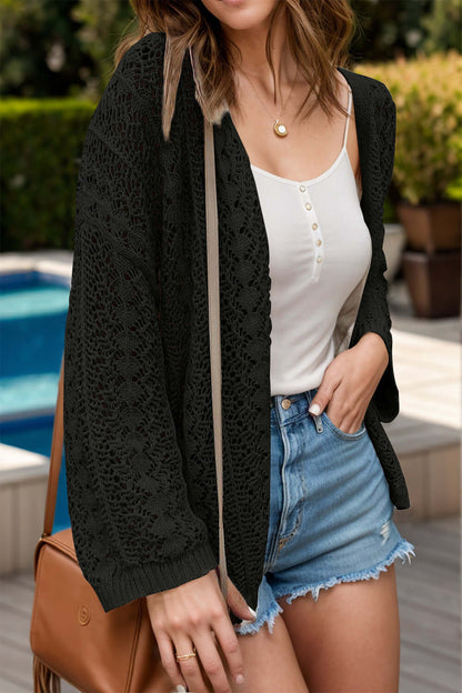 Woman wearing a black openwork open front dropped shoulder cardigan, white tank top, denim shorts, and a brown handbag by the pool.