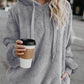 Stylish woman in a cozy gray teddy hoodie, holding a coffee cup, standing on a city street.