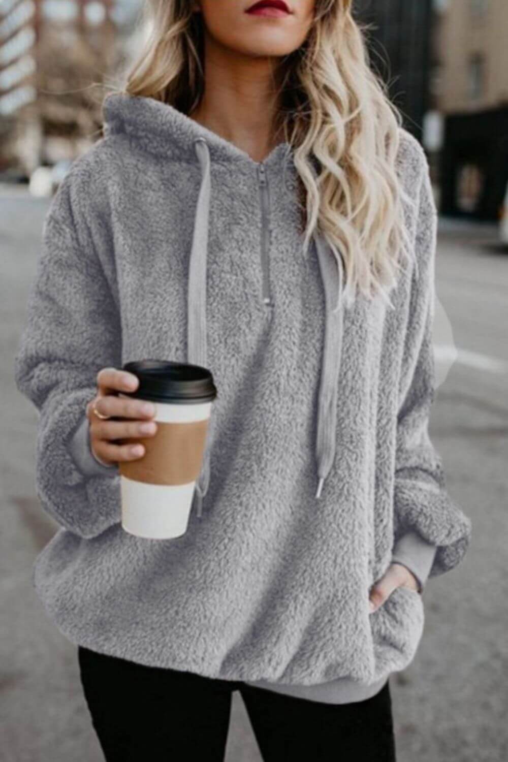 Stylish woman in a cozy gray teddy hoodie, holding a coffee cup, standing on a city street.