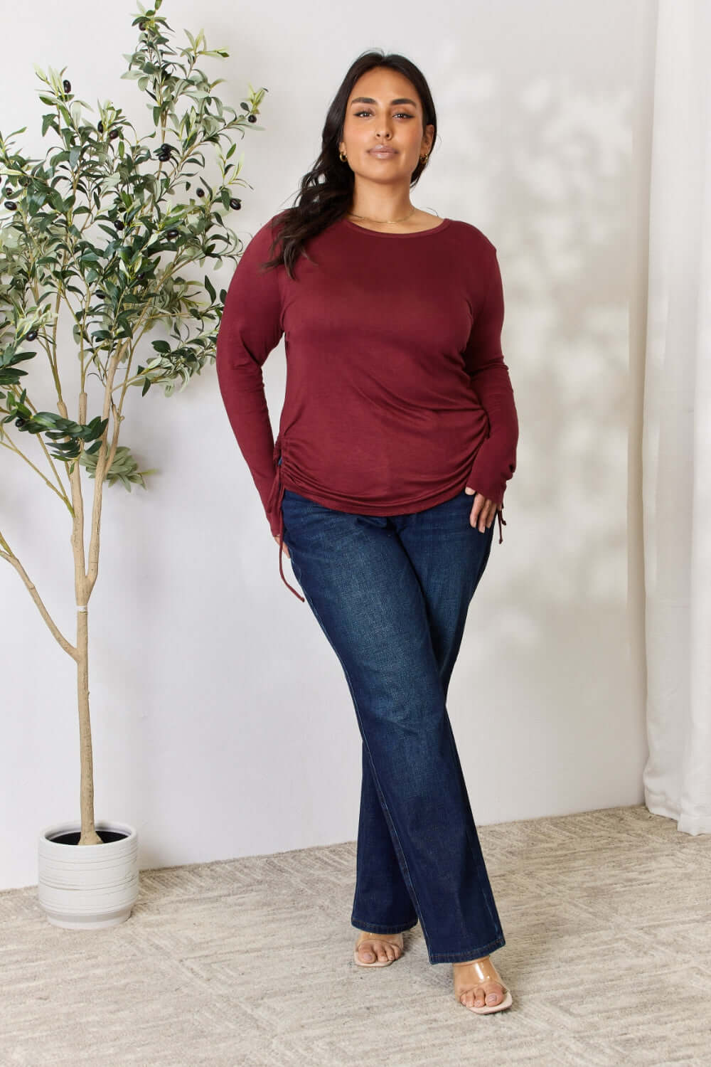 Curvy model wearing a maroon drawstring round neck long sleeve top with jeans, standing next to a plant in a stylish indoor setting.