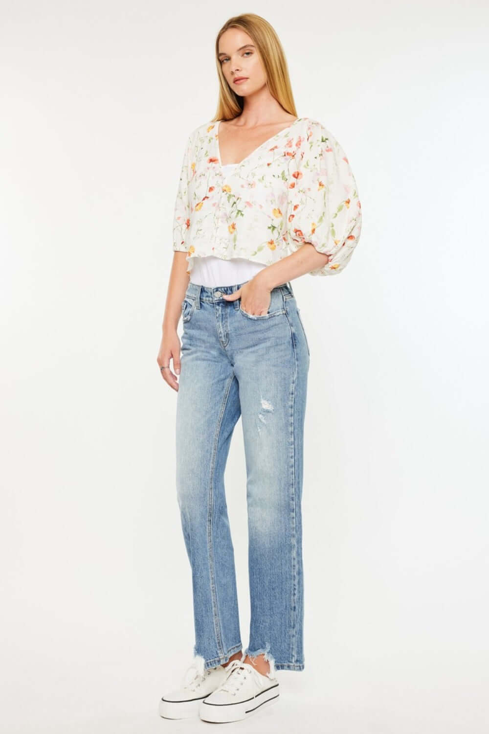 Woman wearing Kancan mid-rise frayed hem straight jeans in medium wash with a floral blouse and white sneakers.