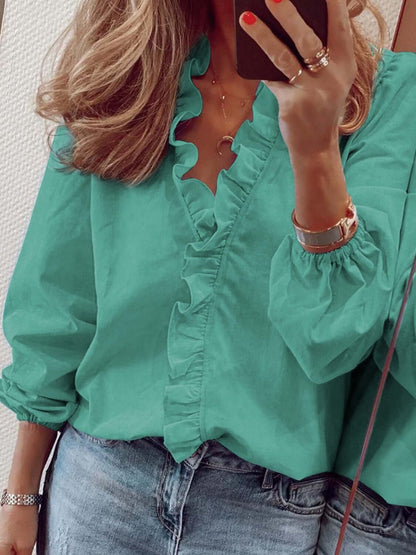 Woman wearing a green ruffled V-neck long sleeve blouse with jeans in a mirror selfie.