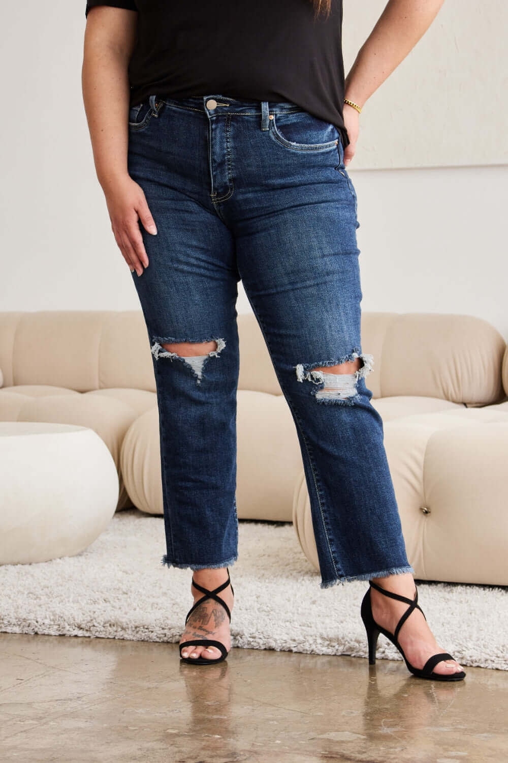 Woman wearing RFM Jeans Crop Dylan full size tummy control distressed high waist raw hem jeans, standing in stylish living room.