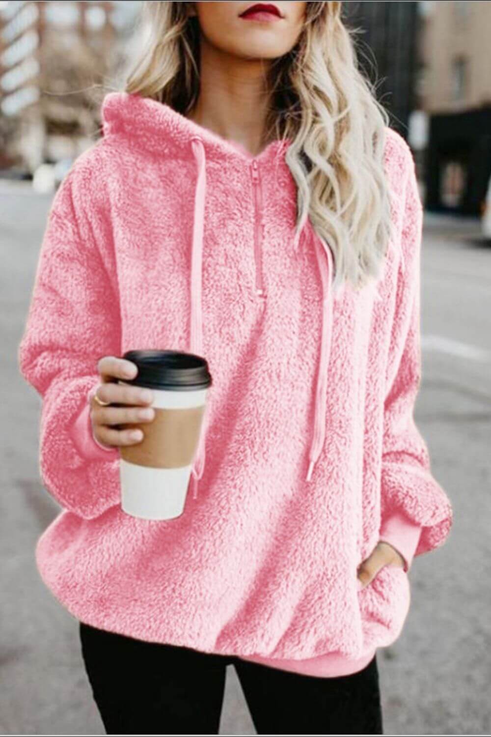 Model wearing a pink Bella Road quarter-zip drawstring teddy hoodie, holding a coffee cup on a city street.