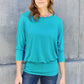 Woman wearing a teal round neck batwing sleeve top paired with jeans, standing against a concrete wall.