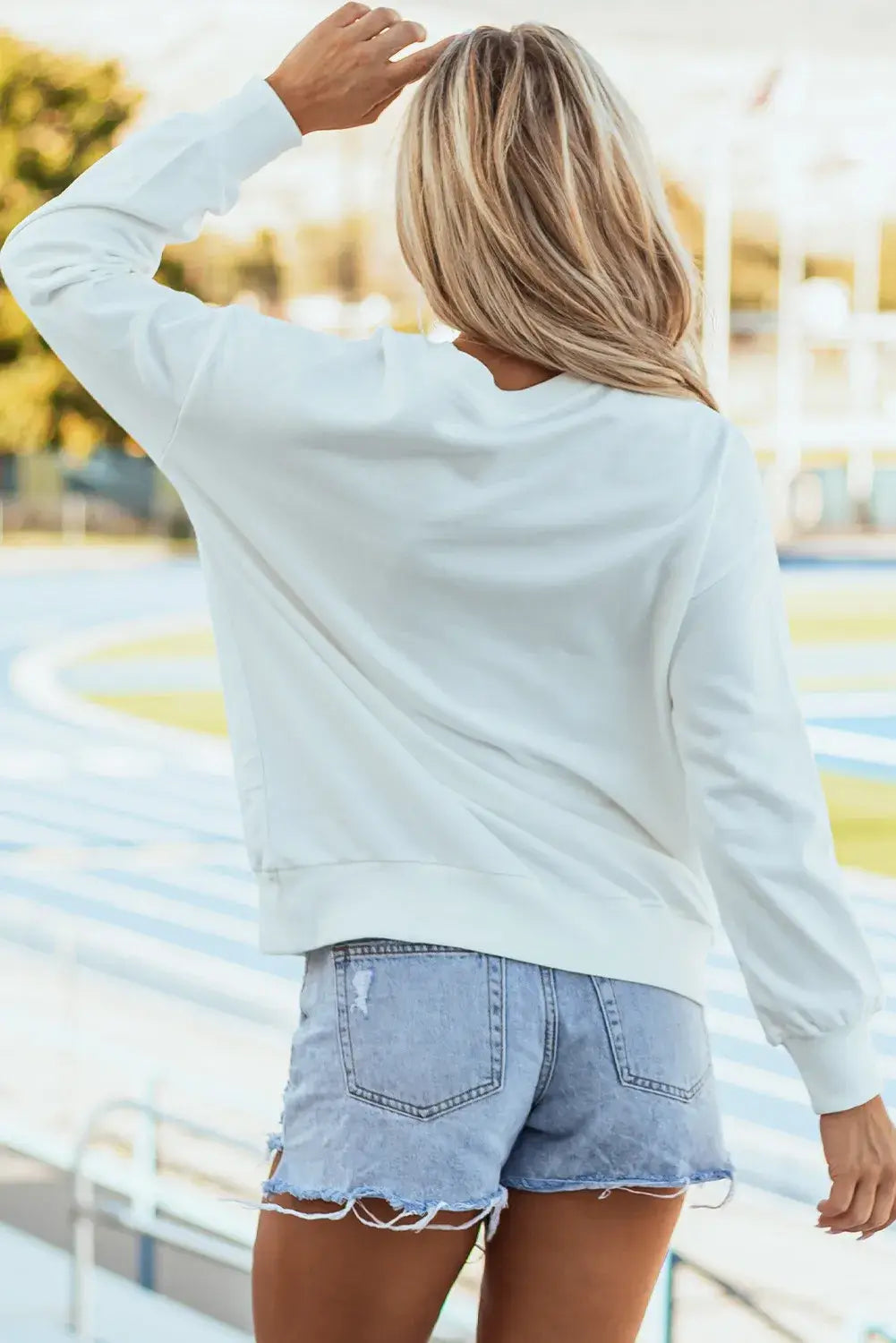 Woman wearing Bella Road Game Day Round Neck Long Sleeve Sweatshirt in white, standing near a sports stadium