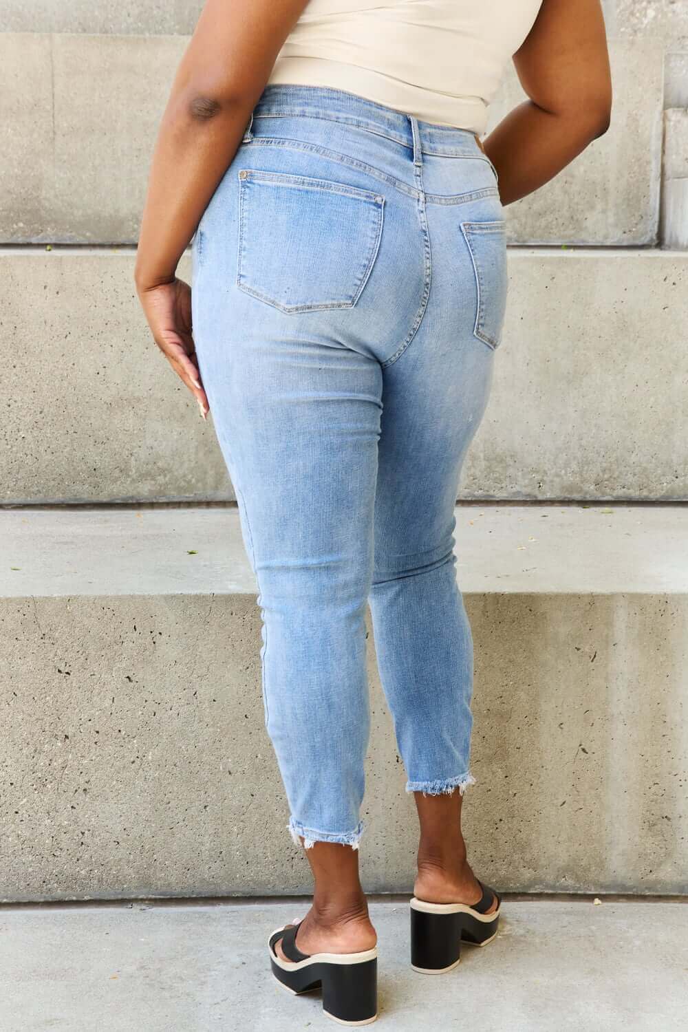 Woman wearing Judy Blue Button Fly Raw Hem Jeans, showcasing back pockets and raw hem on light blue denim.