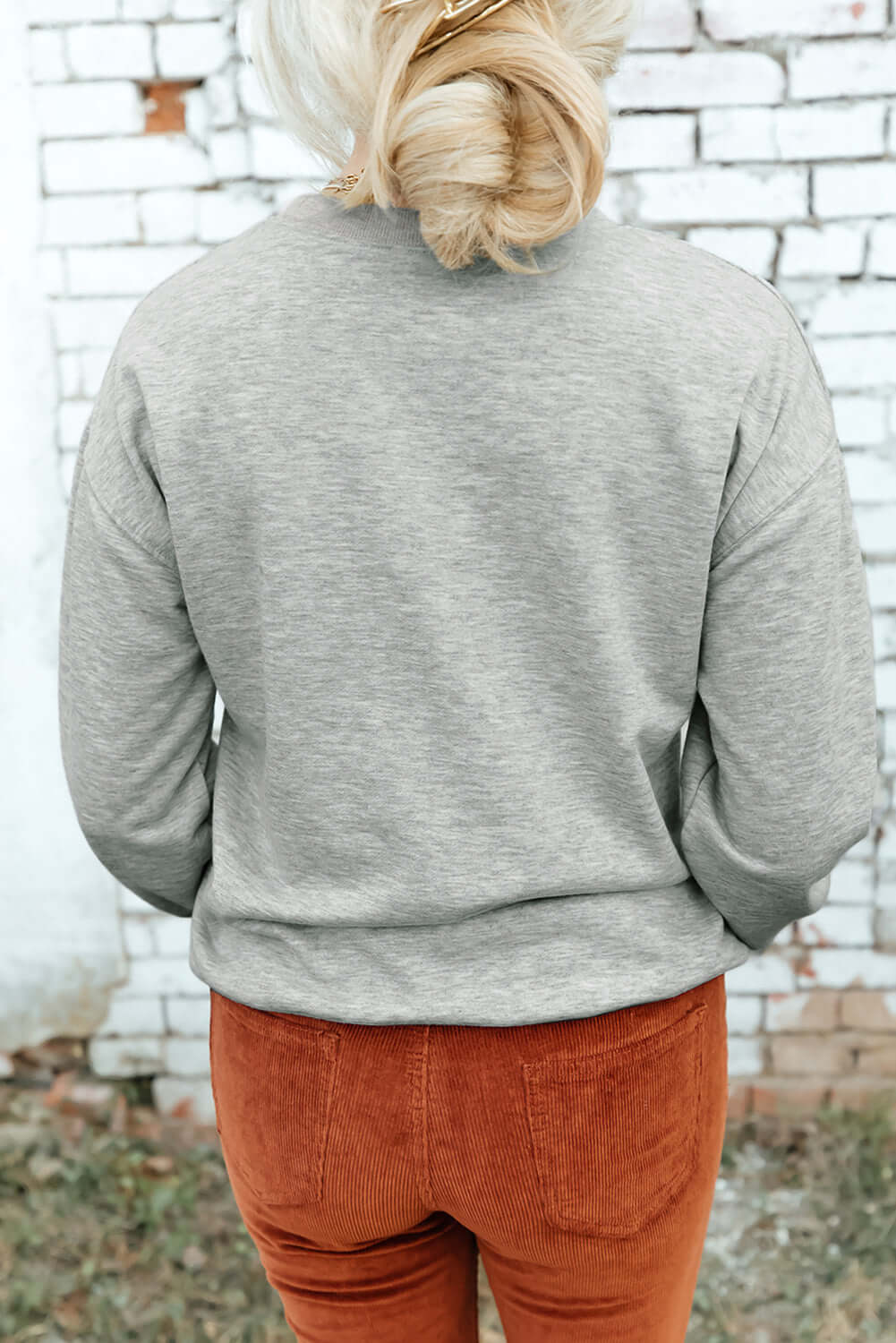 Back view of woman wearing Bella Road Game Day Round Neck Long Sleeve Sweatshirt with orange pants against a white brick wall.