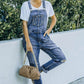 Woman in white tee and pocketed distressed denim overalls with a brown purse, standing outdoors against a white wall with greenery.