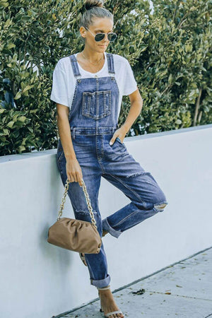 Woman in white tee and pocketed distressed denim overalls with a brown purse, standing outdoors against a white wall with greenery.