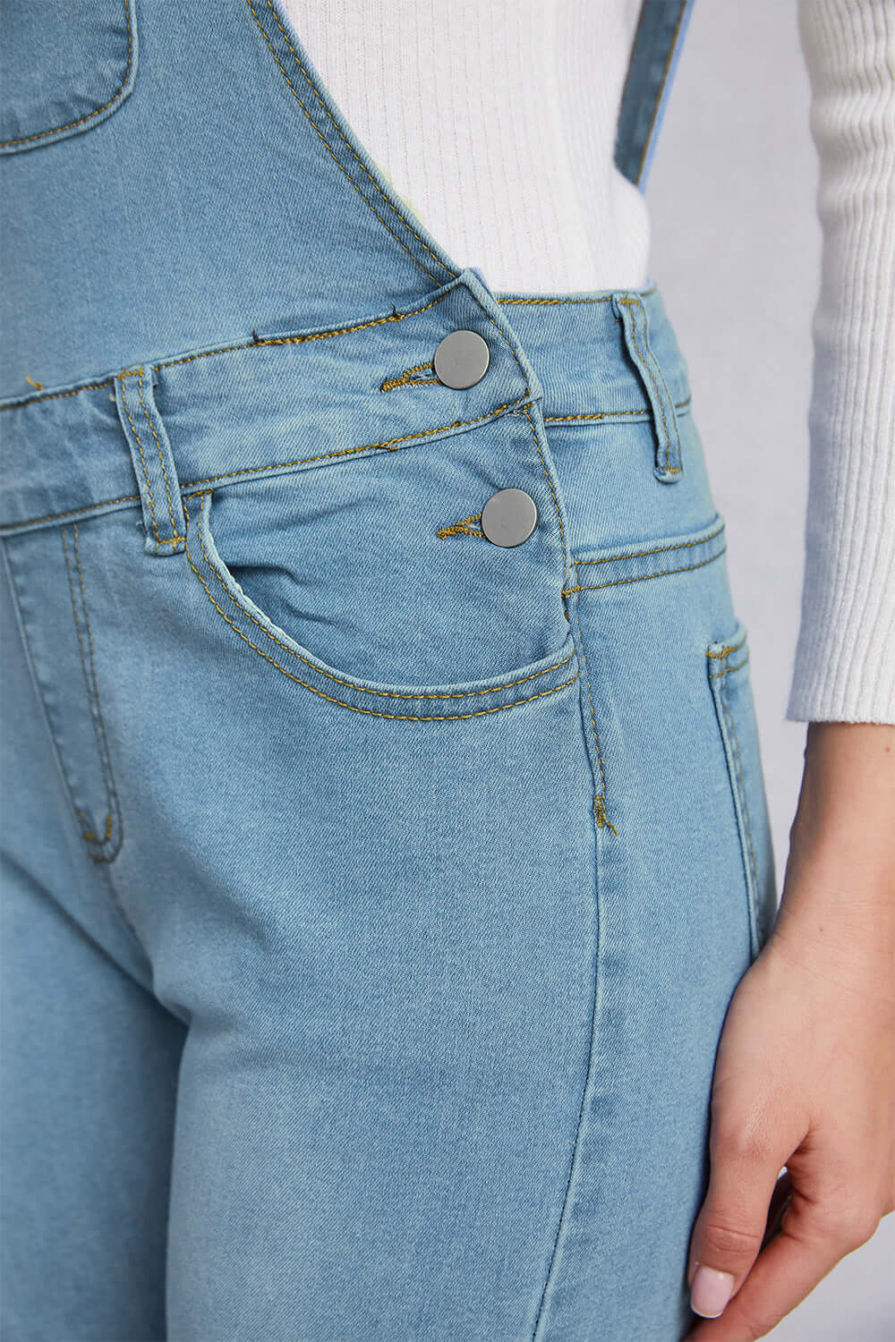 Close-up of distressed washed denim overalls with pockets and button details, featuring a mid waist and slightly stretchy material.