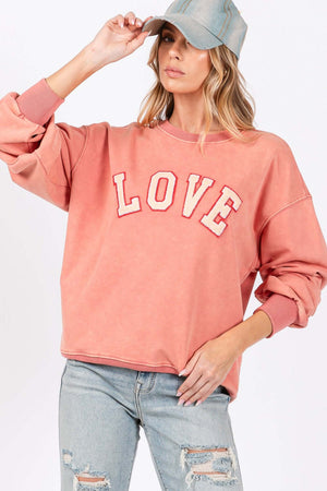 Woman wearing LOVE Path Applique Drop Shoulder Sweatshirt with round neckline and ribbed hems, paired with distressed jeans and a hat