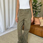 Model wearing RFM Full Size High Rise Garment Dye Wide Leg Jeans in olive green, paired with a white tank top and white shoes.