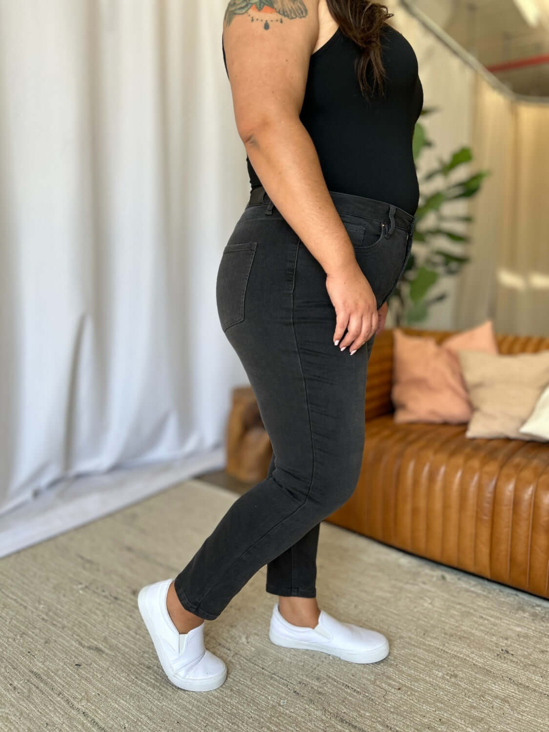 Woman wearing RFM Full Size High Rise Tummy Control Skinny Jeans in dark color, standing indoors near a sofa.