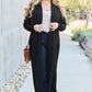 Woman wearing black open front long sleeve cover up, styled with dark jeans, light top, sunglasses, and brown crossbody bag, posing outdoors.