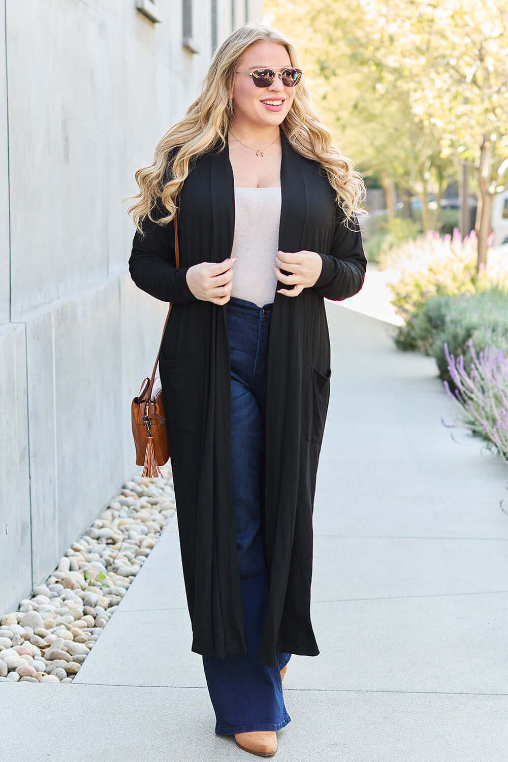 Woman wearing black open front long sleeve cover up, styled with dark jeans, light top, sunglasses, and brown crossbody bag, posing outdoors.