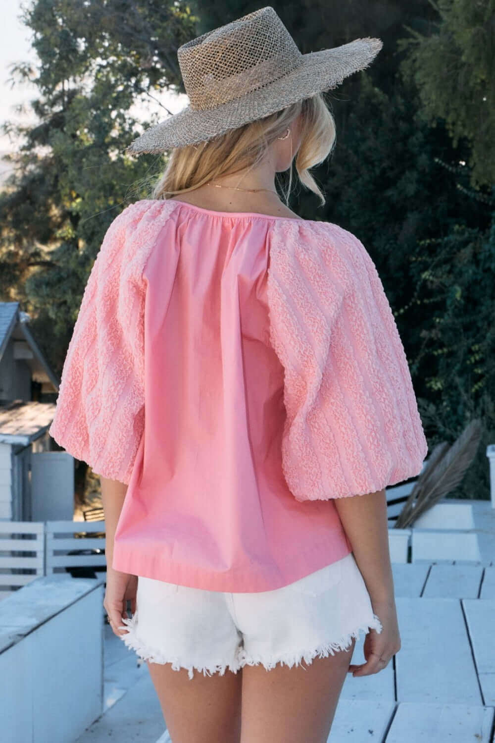 Woman wearing pink poplin bubble sleeve blouse and white shorts, standing outdoors with straw hat