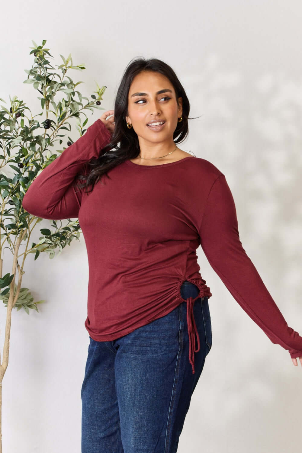 Woman wearing a burgundy Drawstring Round Neck Long Sleeve Top paired with blue jeans, standing in front of a potted plant.