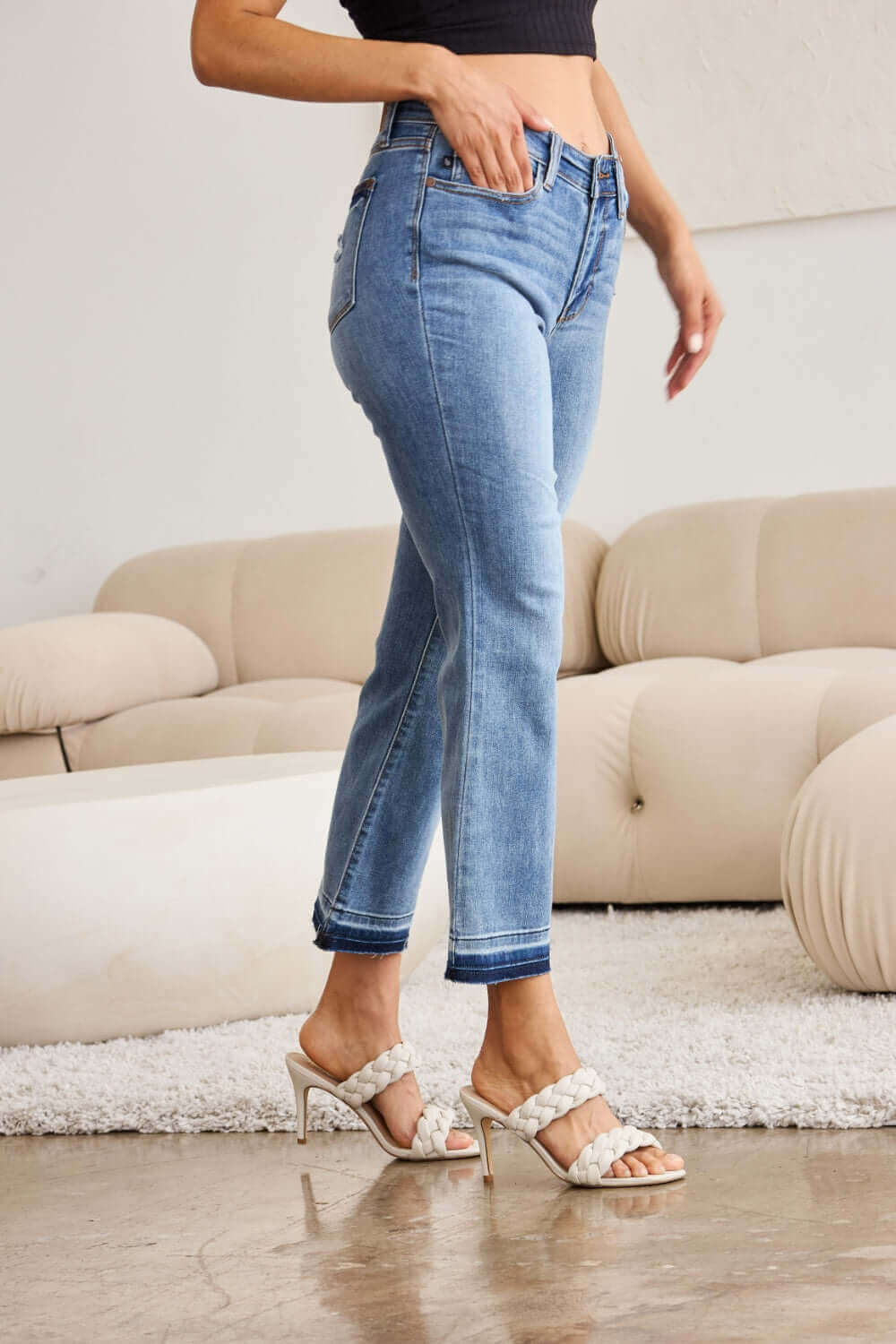 Woman wearing Judy Blue release hem cropped bootcut jeans paired with white heels in a stylish living room setting.