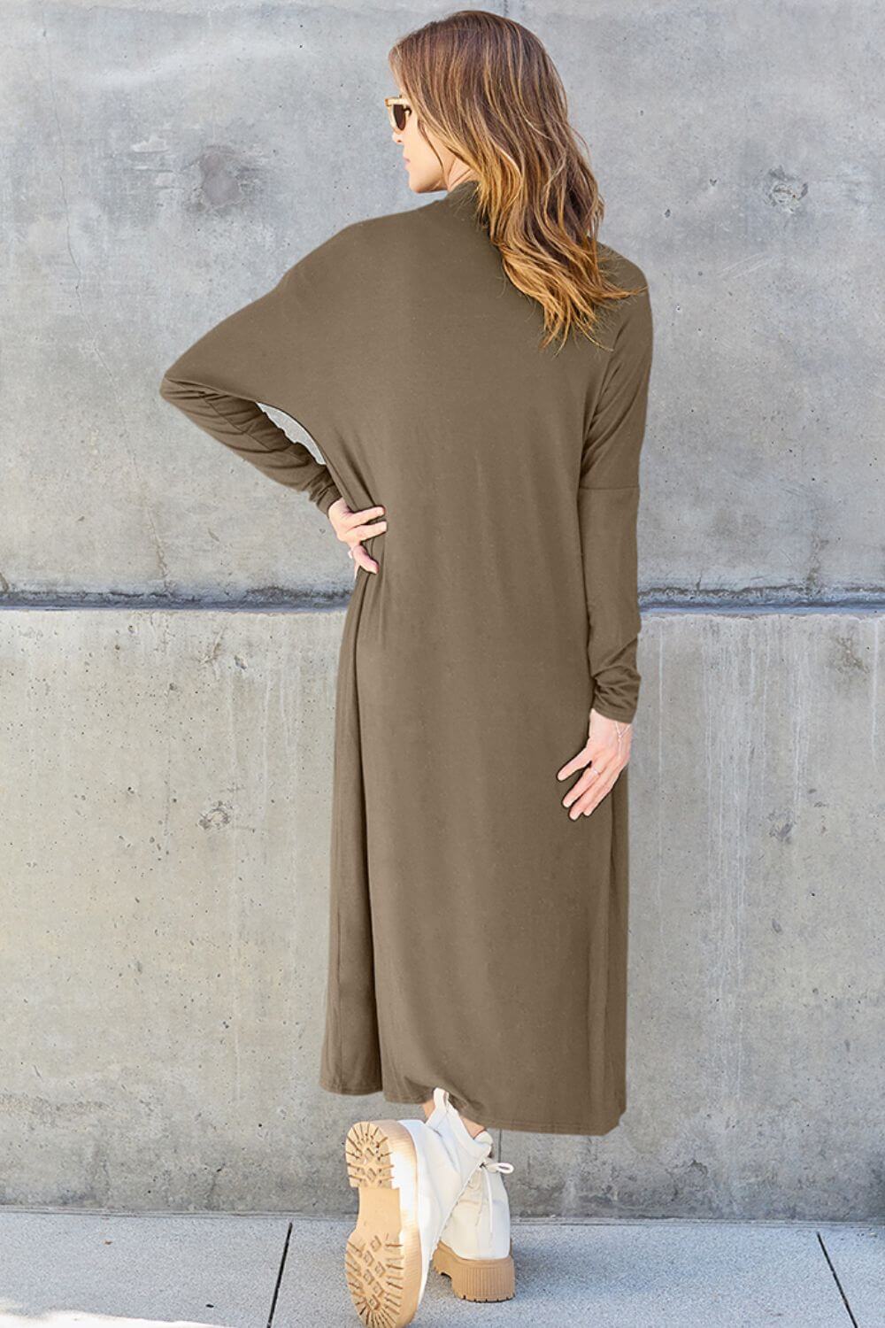 Woman wearing an open front, long sleeve cover up in olive green, showing back view, against a concrete wall.