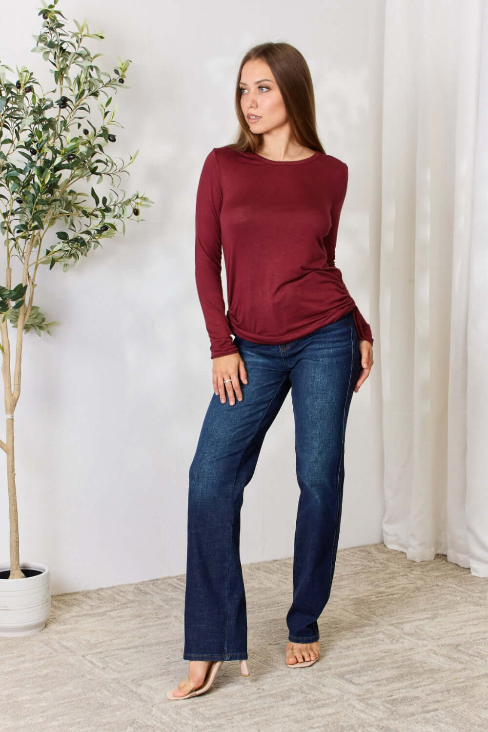 Woman wearing a burgundy drawstring round neck long sleeve top paired with blue jeans, posing in a stylish indoor setting.