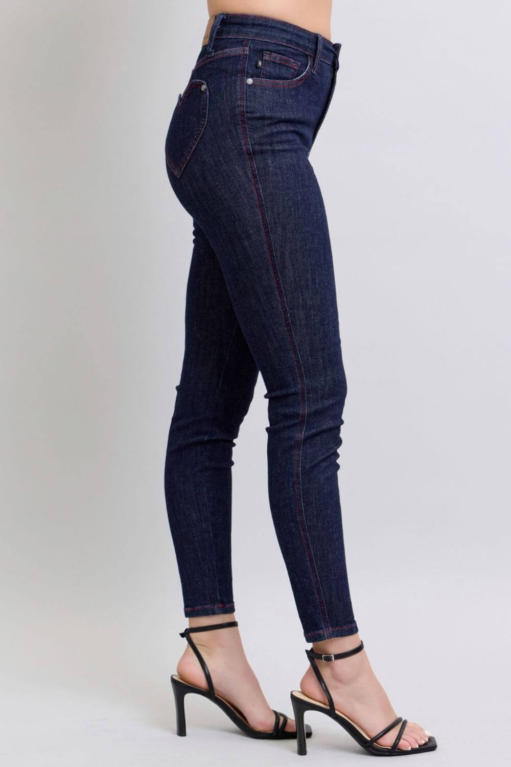 Heart-shaped back pockets skinny jeans in dark denim, side view showing high-rise fit and stylish design, paired with heels.