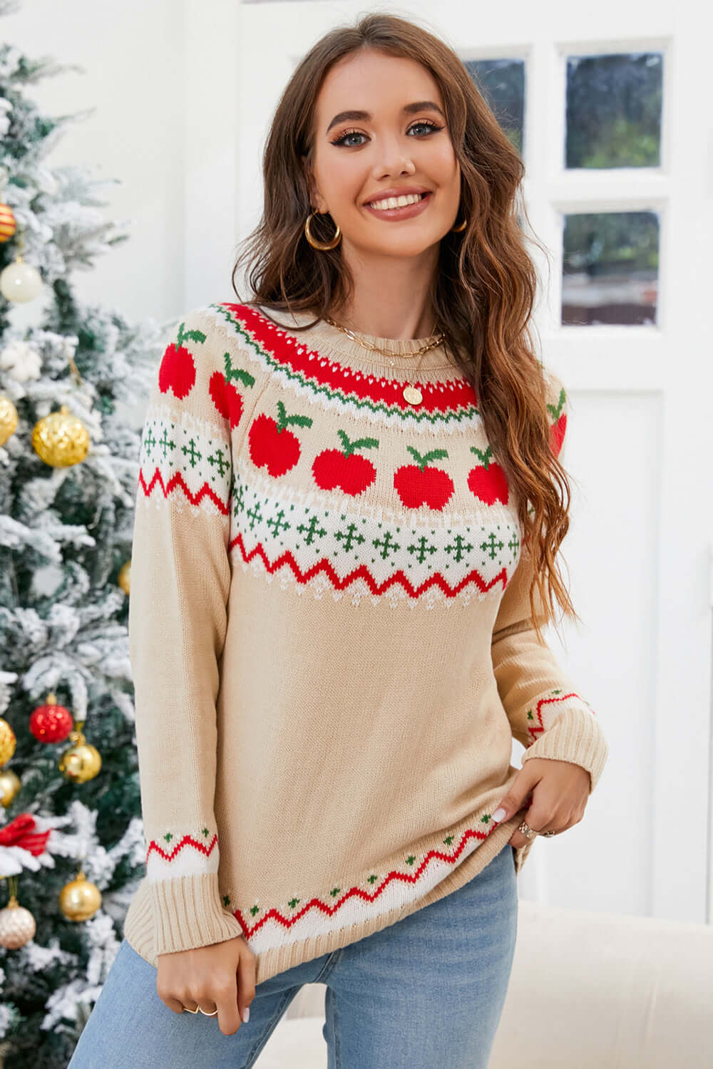 Woman wearing angel wings graphic sweater with apple design, round neck, and long sleeves, standing beside a decorated Christmas tree.