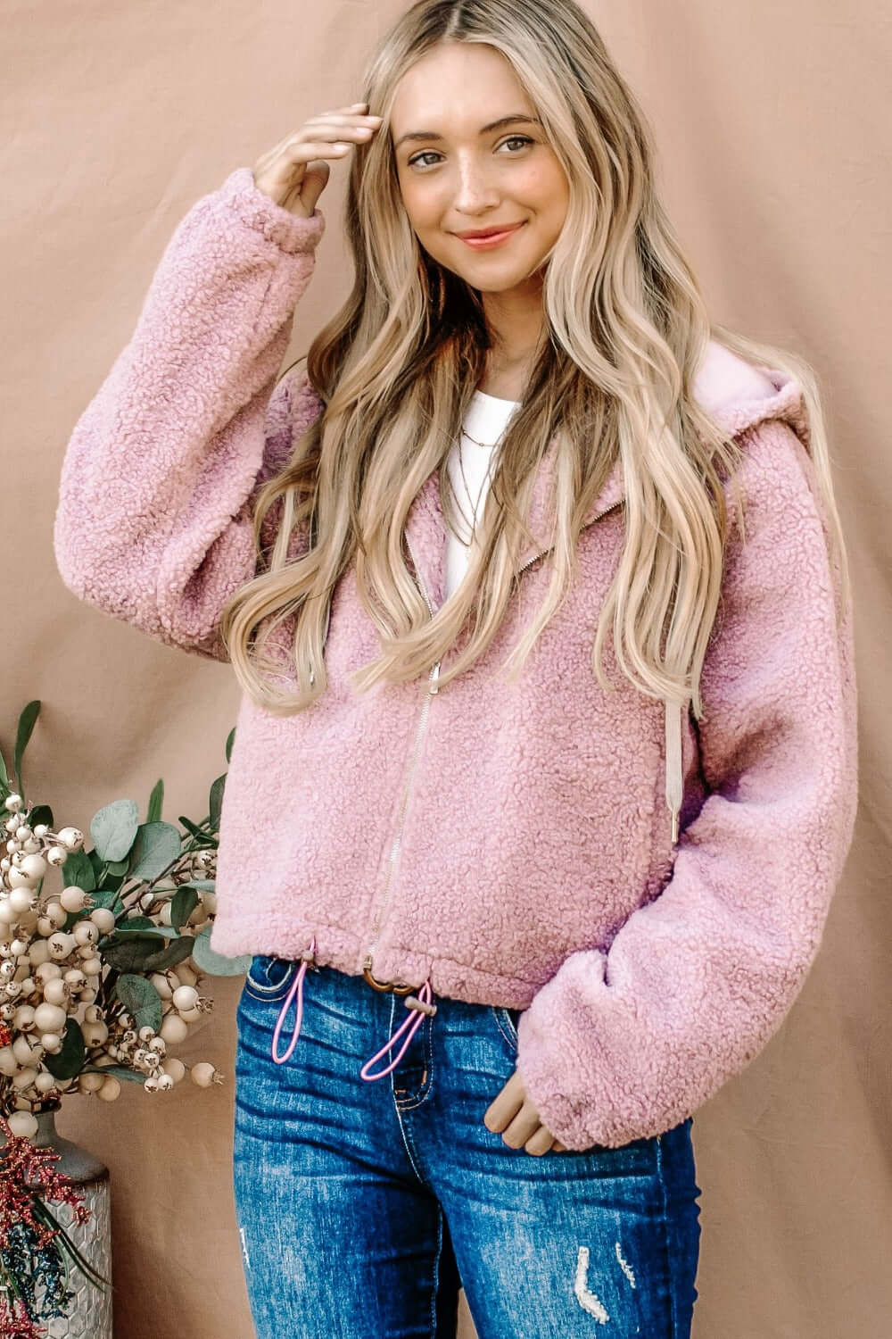 Woman wearing pink Sherpa drawstring cropped hooded jacket with casual jeans against a beige background.