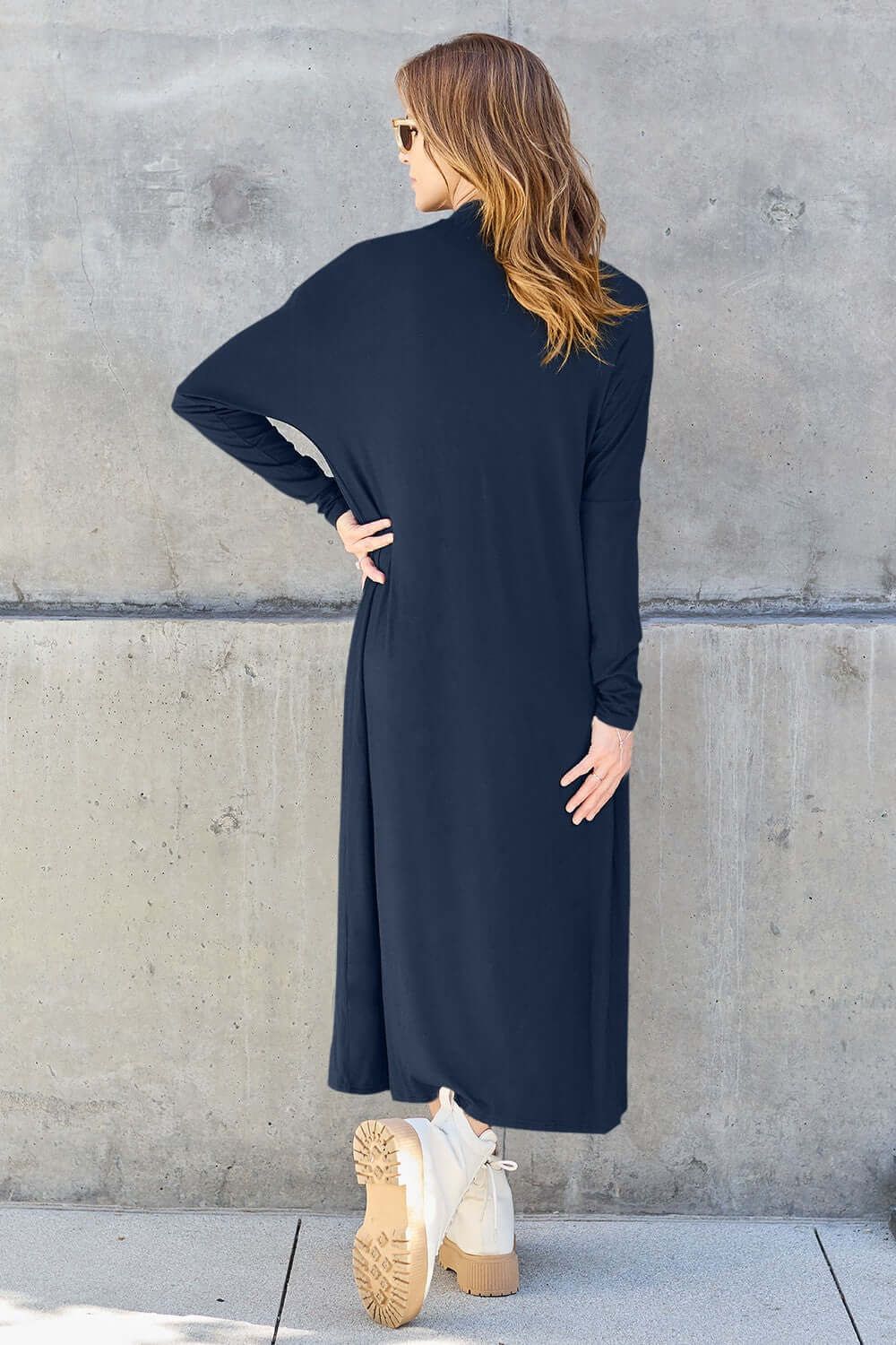 Woman wearing navy open front long sleeve cover up from the back, standing against concrete wall.