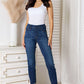 Woman wearing skinny cropped Judy Blue Jeans with a white tank top, styled with heels, standing in a chic, modern room.