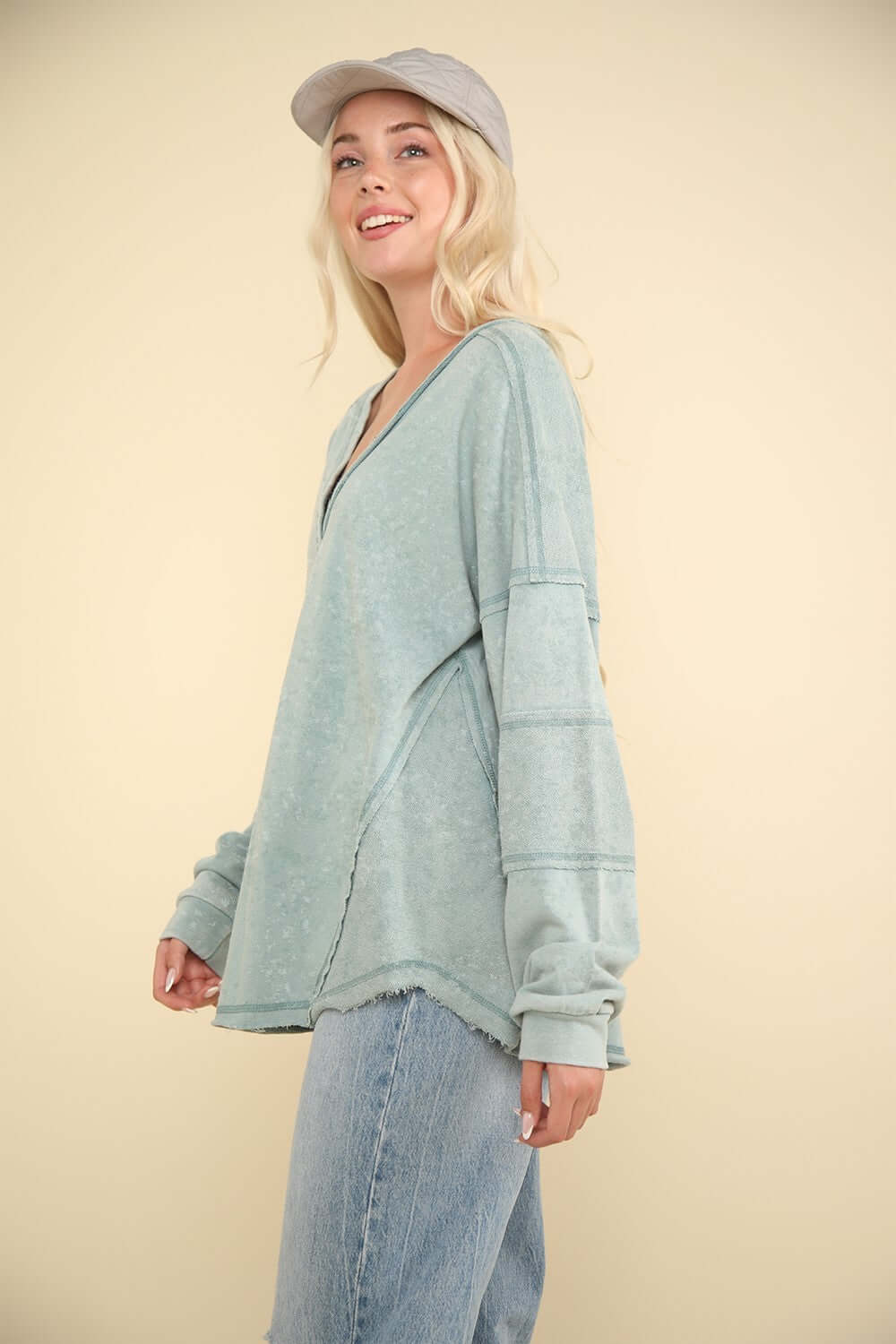 Woman wearing VERY J Washed V-Neck Exposed Seam Knit Top with drop shoulder design and long sleeves, styled with jeans and cap.