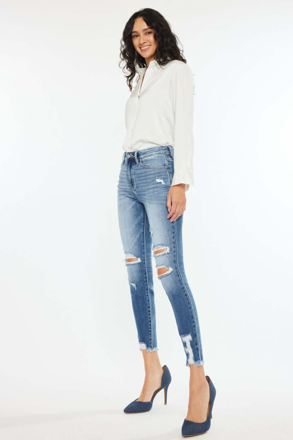 Woman wearing High Rise Frayed Ankle Skinny Jeans in medium wash paired with white top and blue heels.