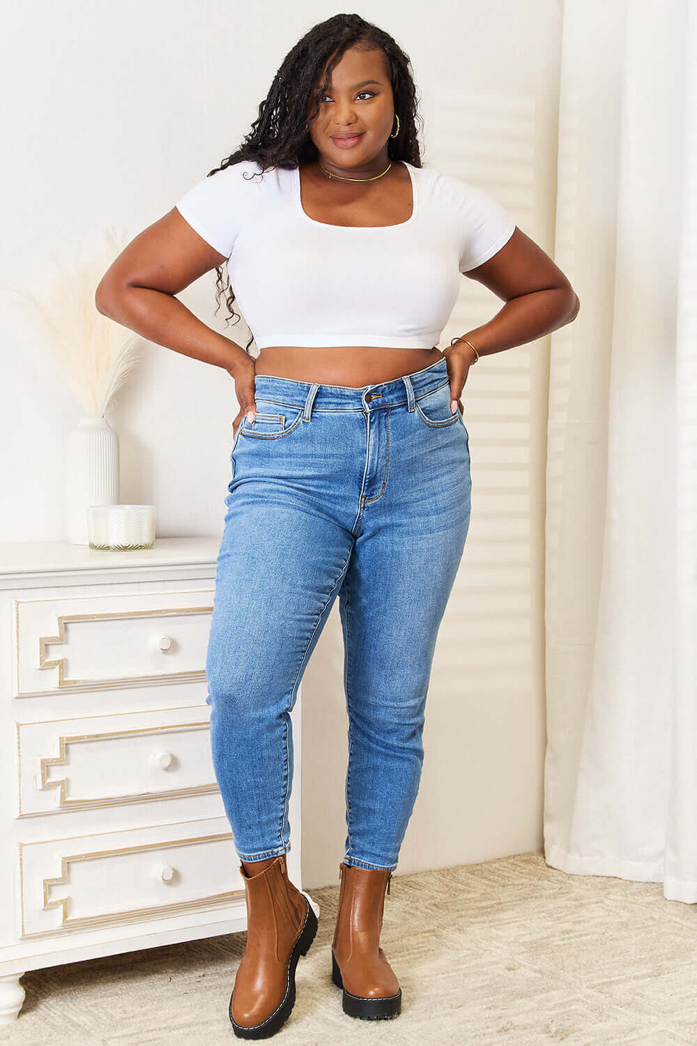 Woman wearing high waist skinny jeans in medium ash wash, paired with a white crop top and brown boots, posing confidently indoors.