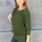 Woman wearing a green round neck batwing sleeve top paired with jeans, standing against a concrete wall.