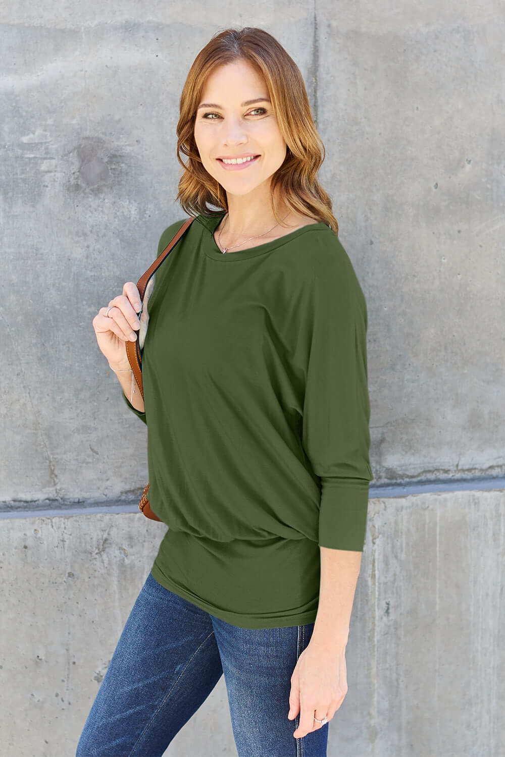 Woman wearing a green round neck batwing sleeve top paired with jeans, standing against a concrete wall.