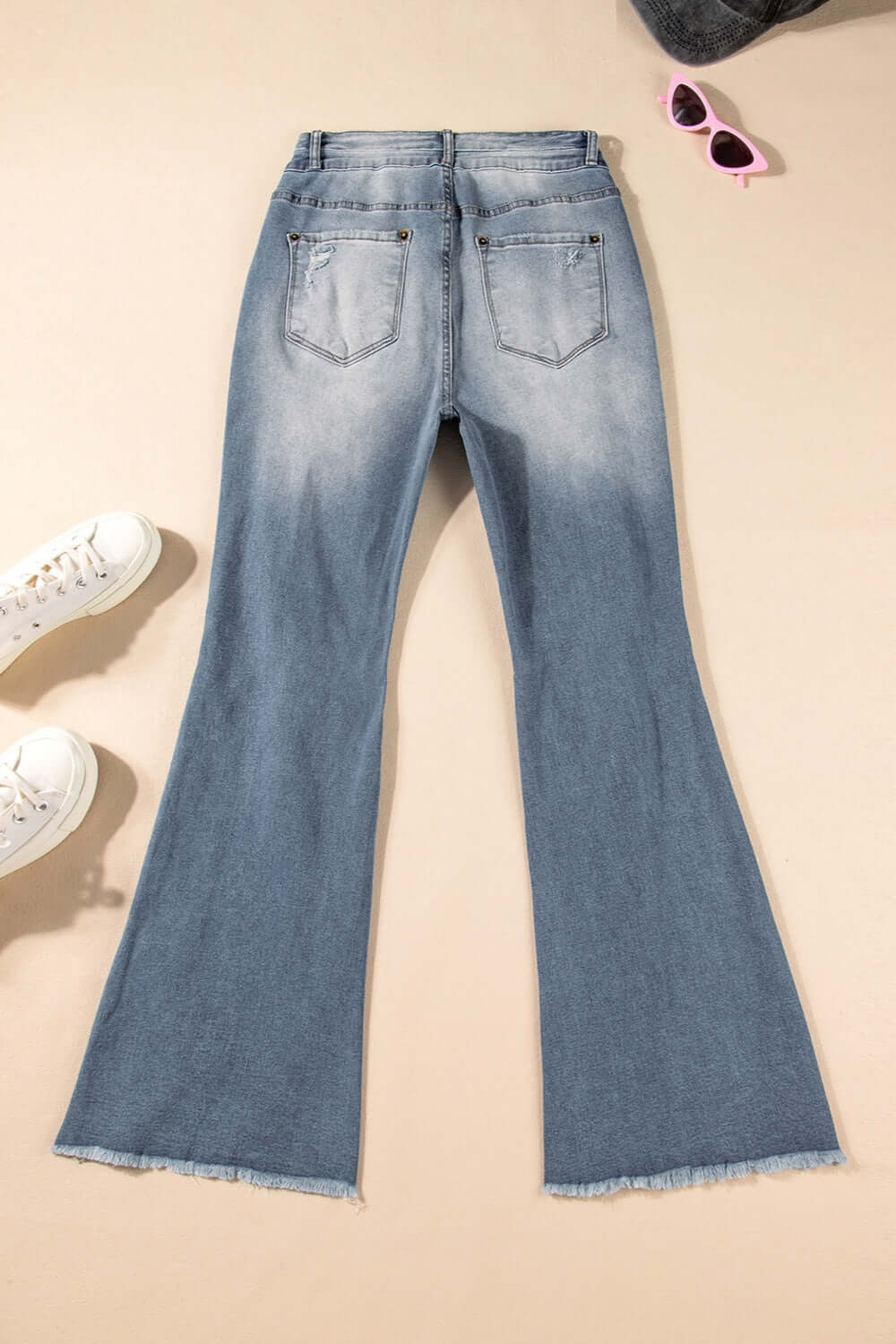 Distressed raw hem bootcut jeans for women with slightly stretchy material laid flat with sunglasses and sneakers accessories.