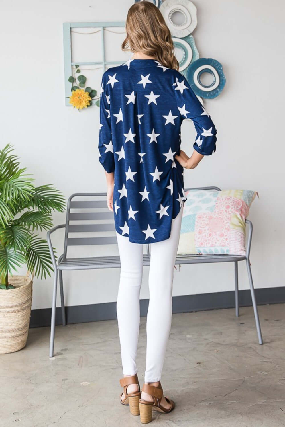 Woman wearing Heimish Roll-Tab Sleeve Star Print Top in navy with white stars, paired with white jeans and sandals, back view.