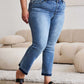 Woman wearing Judy Blue release hem cropped bootcut jeans with black sandals in a contemporary living room setting.