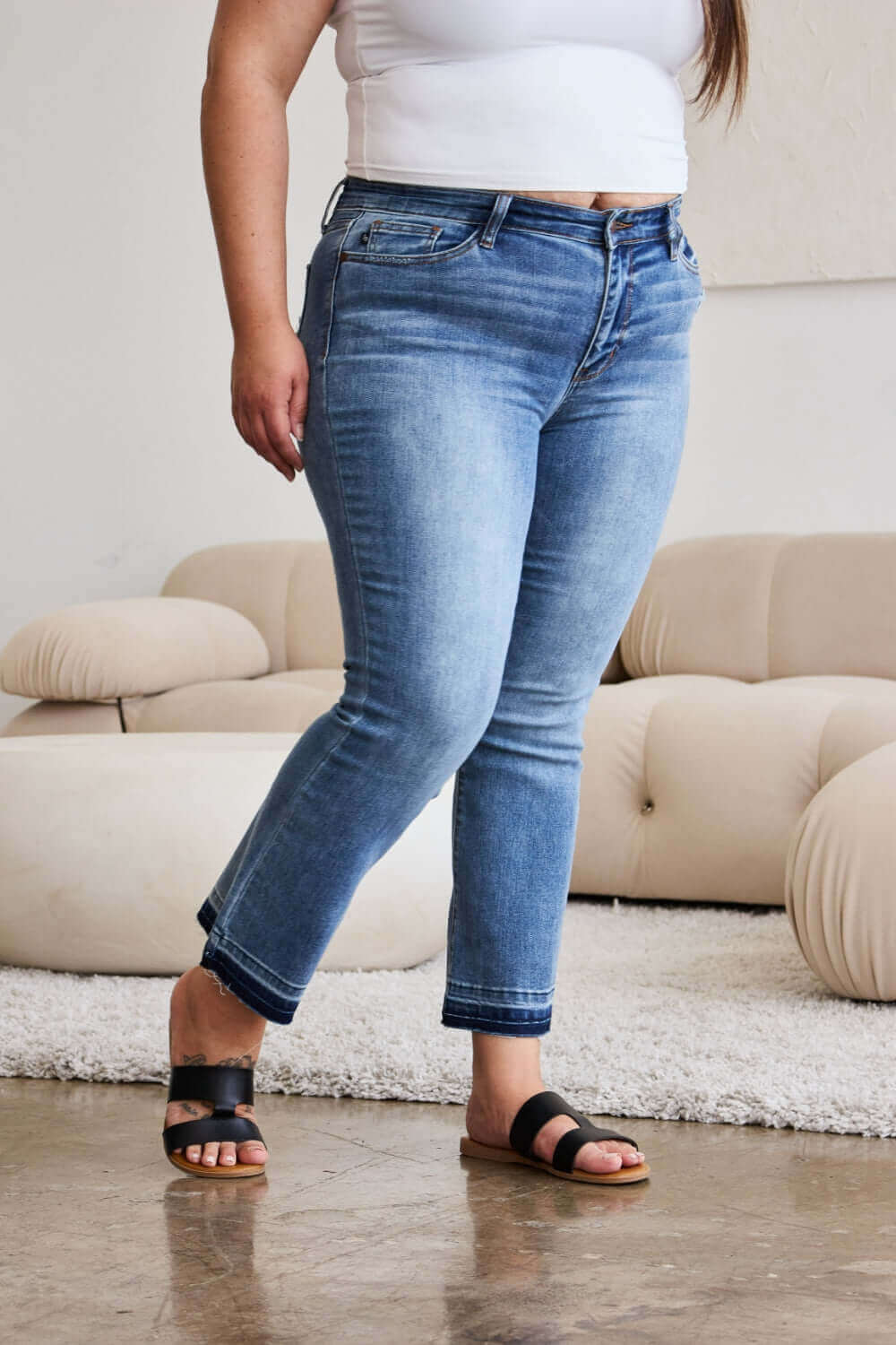 Woman wearing Judy Blue release hem cropped bootcut jeans with black sandals in a contemporary living room setting.