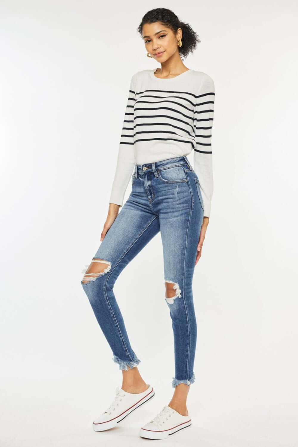 Woman modeling high waist distressed raw hem ankle skinny jeans, paired with a striped sweater and white sneakers. Trendy and edgy look.