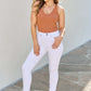 Woman wearing Alyssa High Rise Skinny Jeans in white, paired with a brown tank top and blue heeled sandals, posing outdoors.