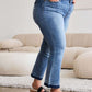 Woman wearing Judy Blue release hem cropped bootcut jeans and black sandals standing in a modern living room.