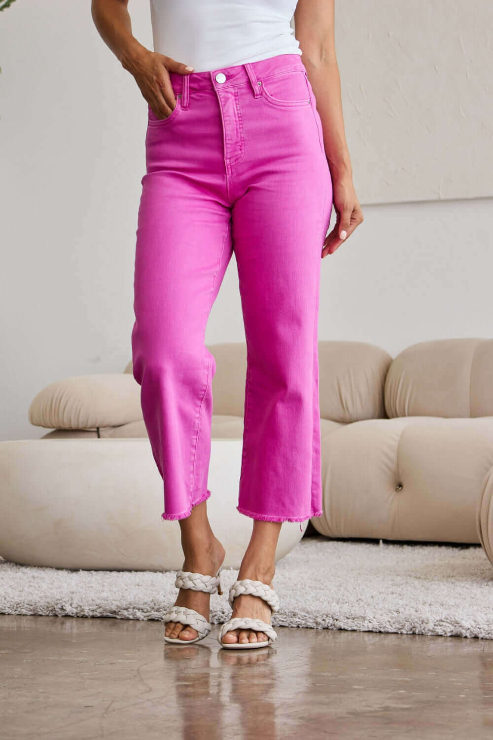 Model wearing Crop Chloe Full Size Tummy Control High Waist Raw Hem Jeans in bright pink, standing in a modern living room. RFM Jeans.