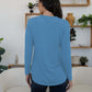 Woman wearing blue FAM-FAM round neck long sleeve t-shirt, showcasing back view in a stylish room with plants and shelves.