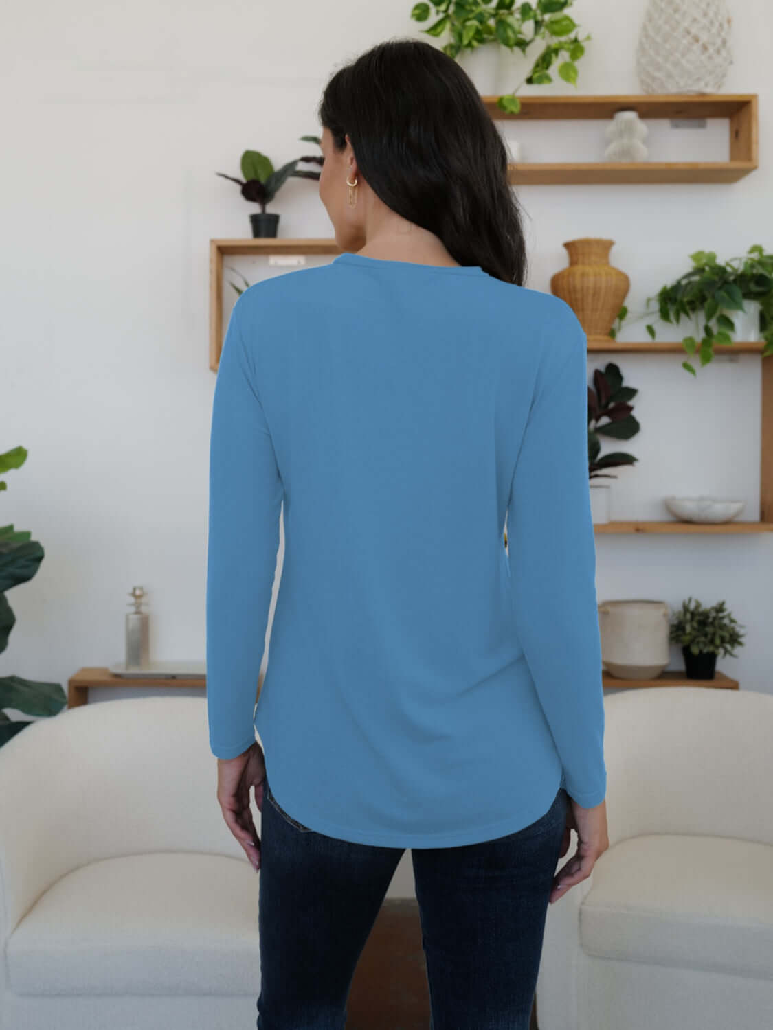 Woman wearing blue FAM-FAM round neck long sleeve t-shirt, showcasing back view in a stylish room with plants and shelves.