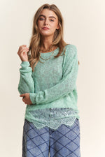 Woman wearing J.NNA lace detail long sleeve knit top in mint green, featuring intricate lace accents, paired with casual blue trousers.