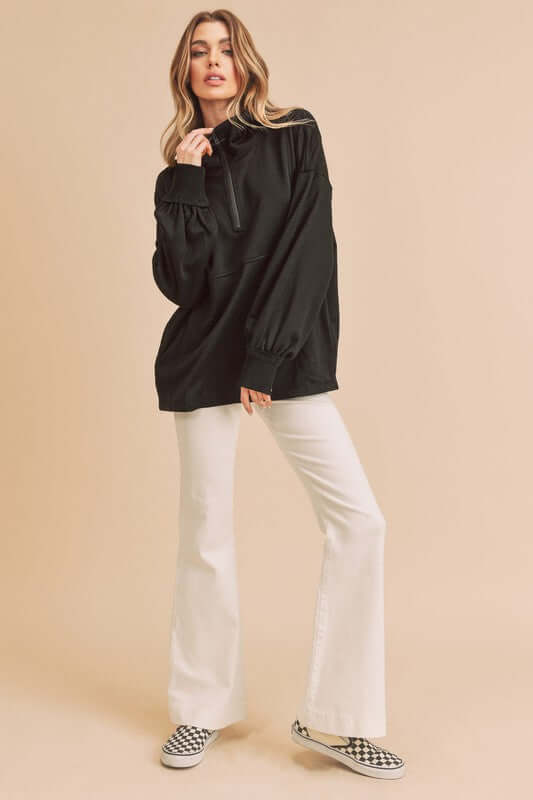 Woman modeling Aemi+Co exposed seam half zip drop shoulder sweatshirt with flared pants and checkered shoes on a neutral background.