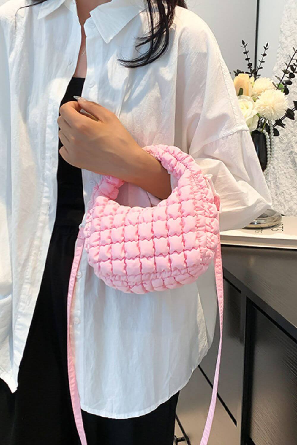 Woman holding a small pink quilted puffy crossbody bag with a removable strap, styled as a clutch against a white shirt backdrop.