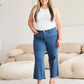 Woman wearing Crop Chloe Full Size Tummy Control High Waist Raw Hem Jeans with white top and sandals in stylish living room